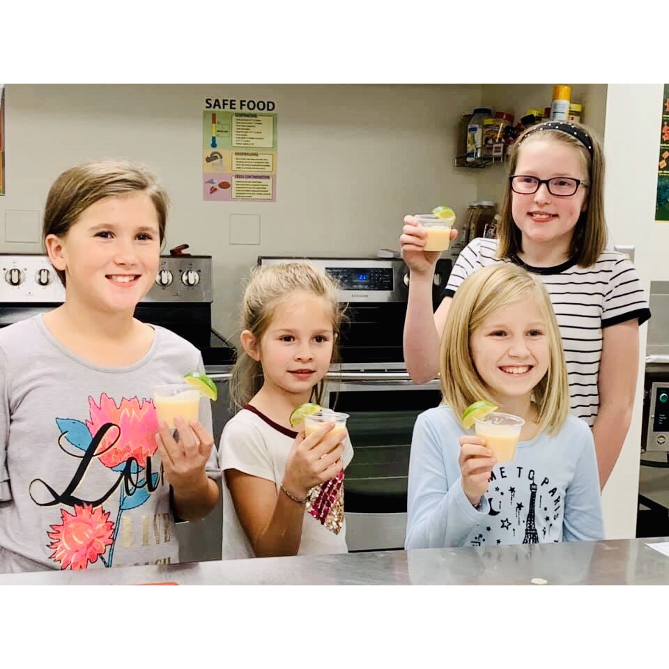 Four 4-H members smile while holding orange smoothies in plastic cups garnished with a lime.